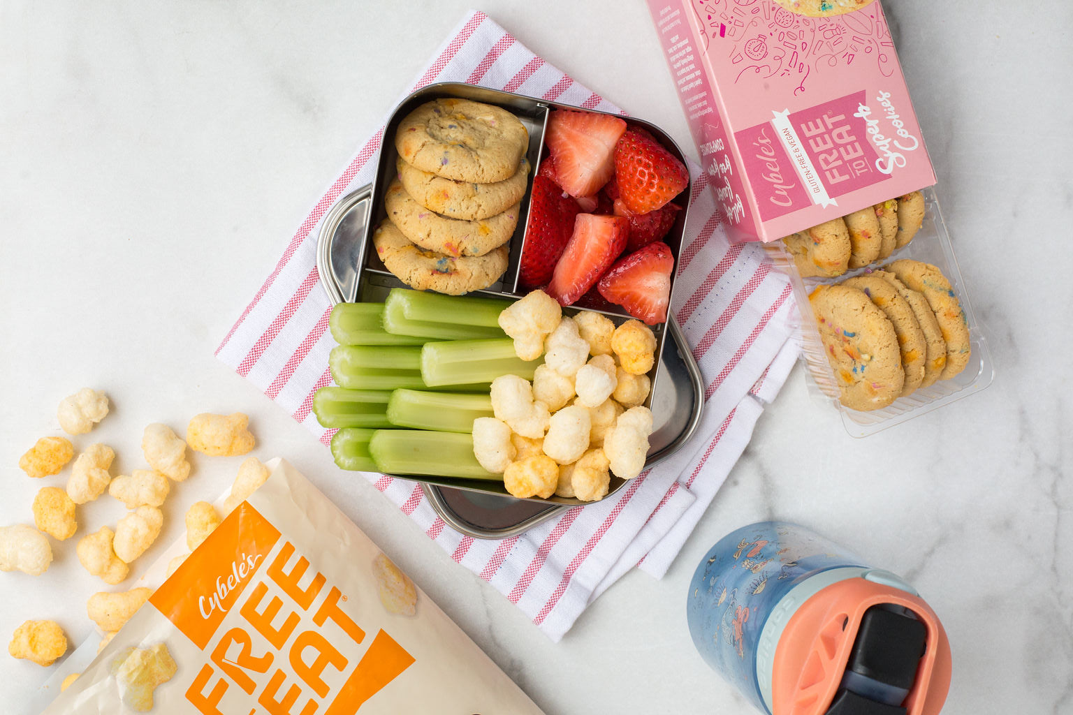 kids bento box on counter with Bag of Cybeles Cheddar Ancient Grain Pops, box of Confetti Cookies and Water bottle. Bento is filled with celery, ancient grain Pops, confetti cookies and strawberries